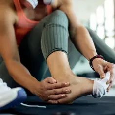 Kobieta masuje bolącą kostkę w siłowni, obok leżą buty sportowe.