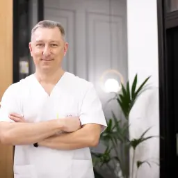 Mężczyzna w białej koszuli lekarskiej ze skrzyżowanymi rękami, uśmiechnięty.