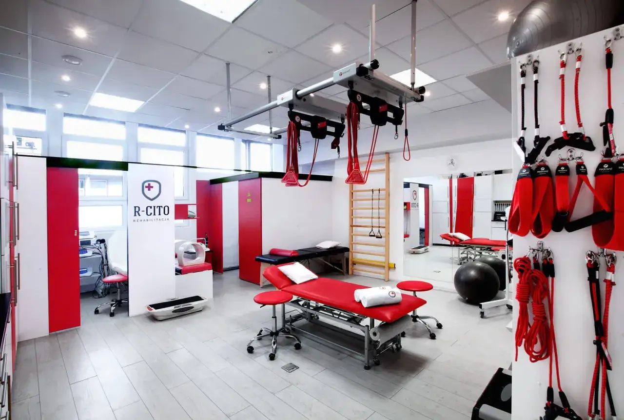 Nowoczesna sala do kinezyterapii z czerwonymi łóżkami rehabilitacyjnymi, drabinką, piłkami i systemem podwieszeń.